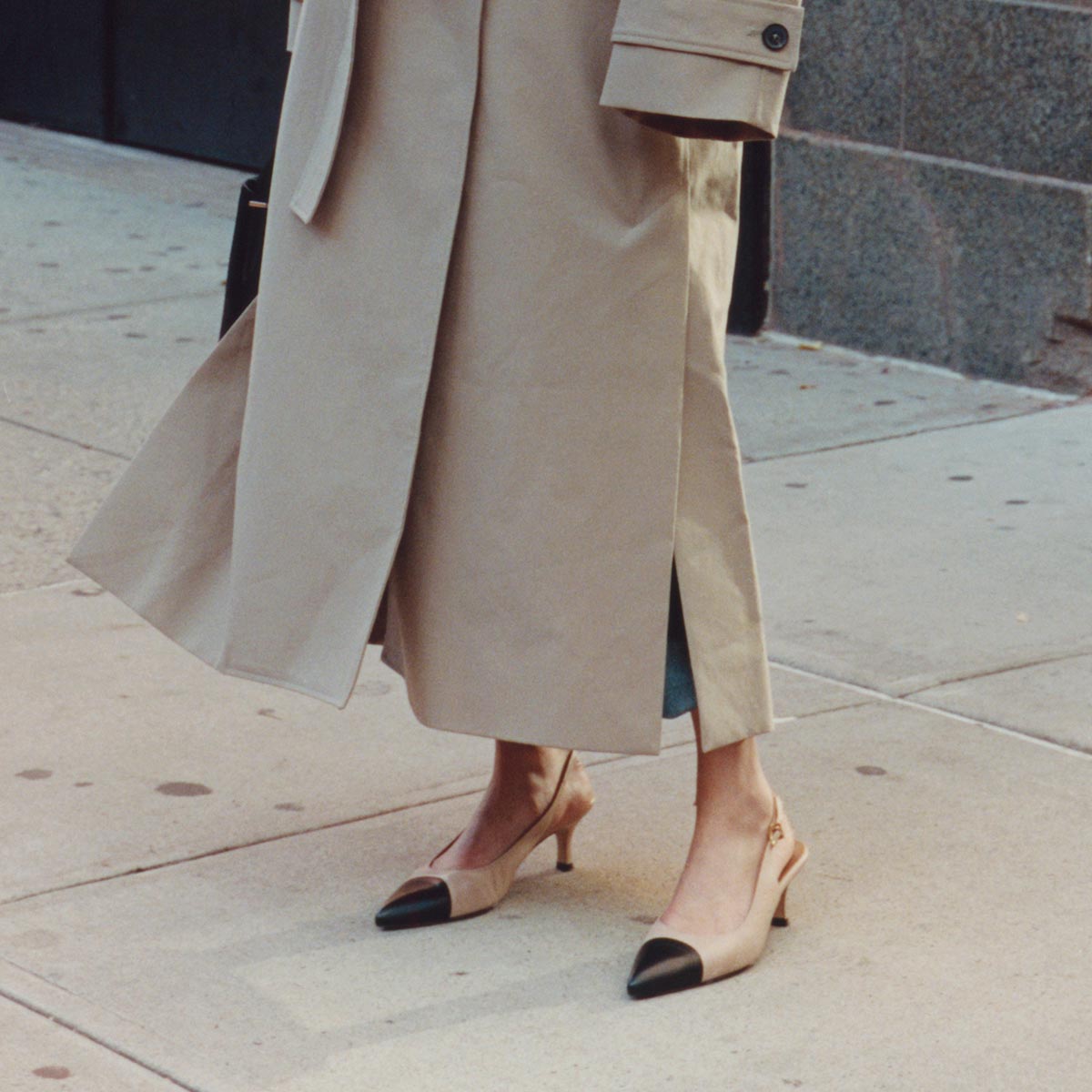 A person in a beige trench coat and Margaux's The Colette - Dune and Black Nappa slingback heels with a micro heel and black snip toe stands on a city sidewalk.