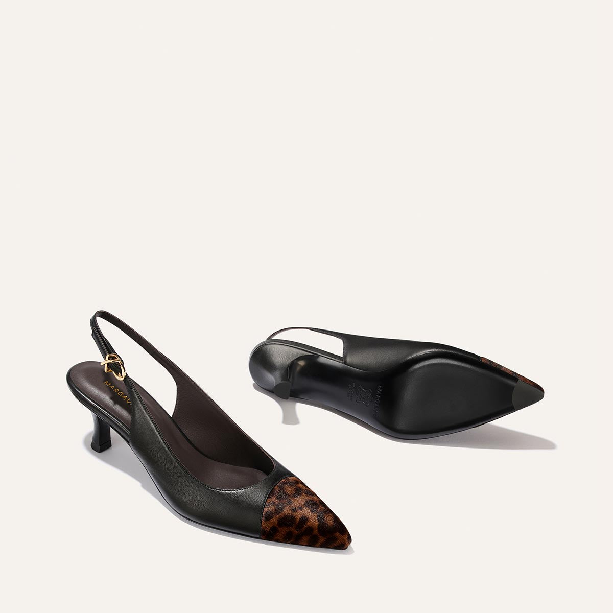 A pair of Margaux’s The Colette heels in black nappa and leopard haircalf, featuring a snip toe and a small micro heel, are displayed on a light background—one standing upright and the other resting on its side.