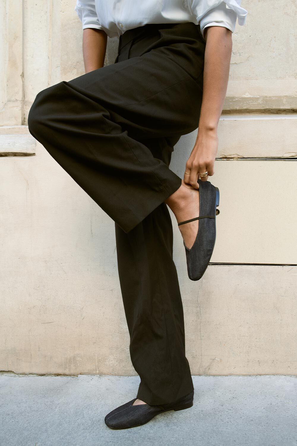 A person dressed in black trousers and a white shirt balances on one leg while adjusting a black flat shoe, standing against a light-colored wall with a concrete sidewalk beneath.