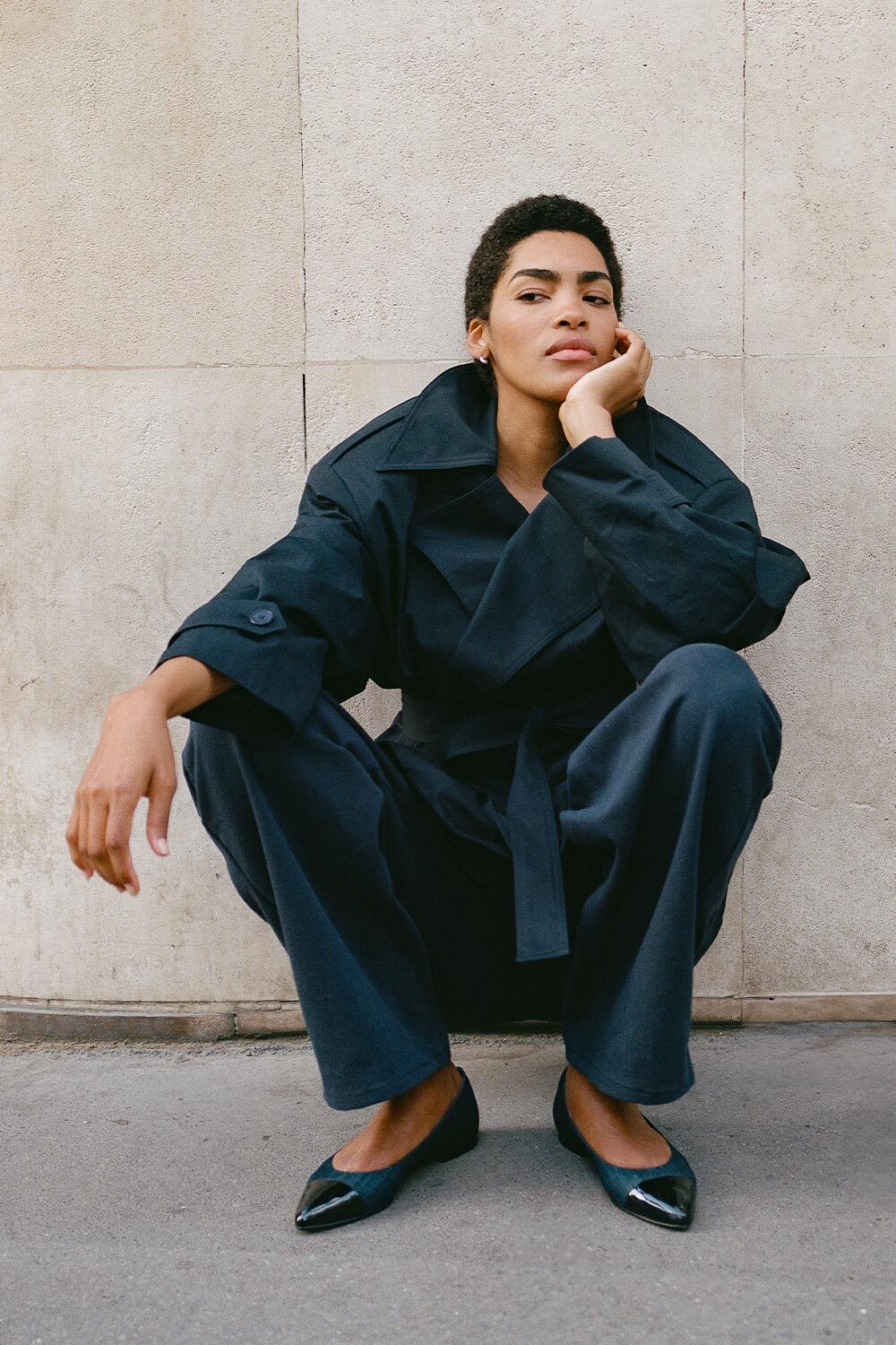 A person with short hair wearing a dark, loose-fitting suit and black pointed shoes squats against a light stone wall, resting their chin on one hand and looking confidently at the camera.
