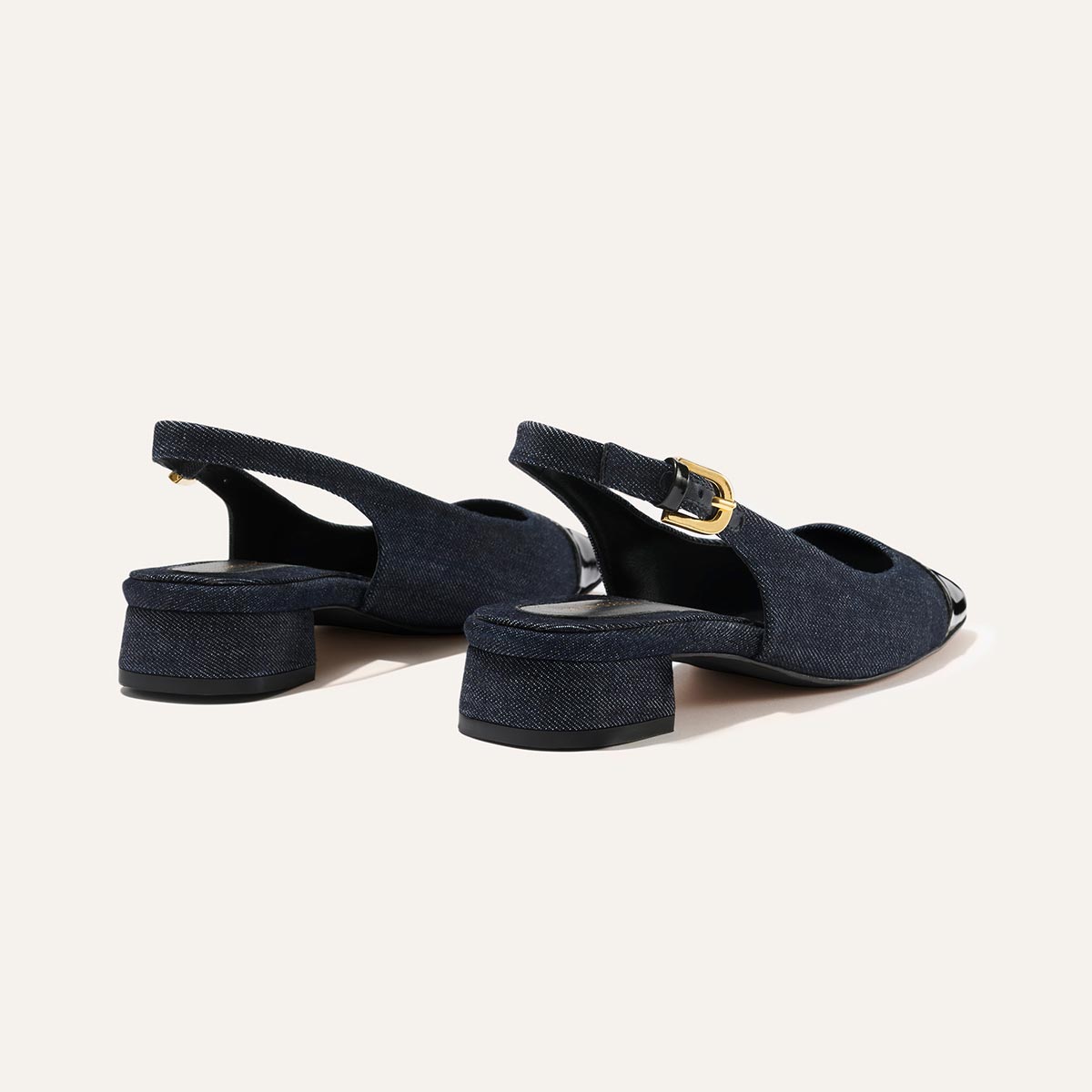 The Darcy Slingback by Margaux, in indigo denim and patent with black capped toes, cutout sides, gold buckle straps, and low block heels, channels ballet pump charm against a plain light background.