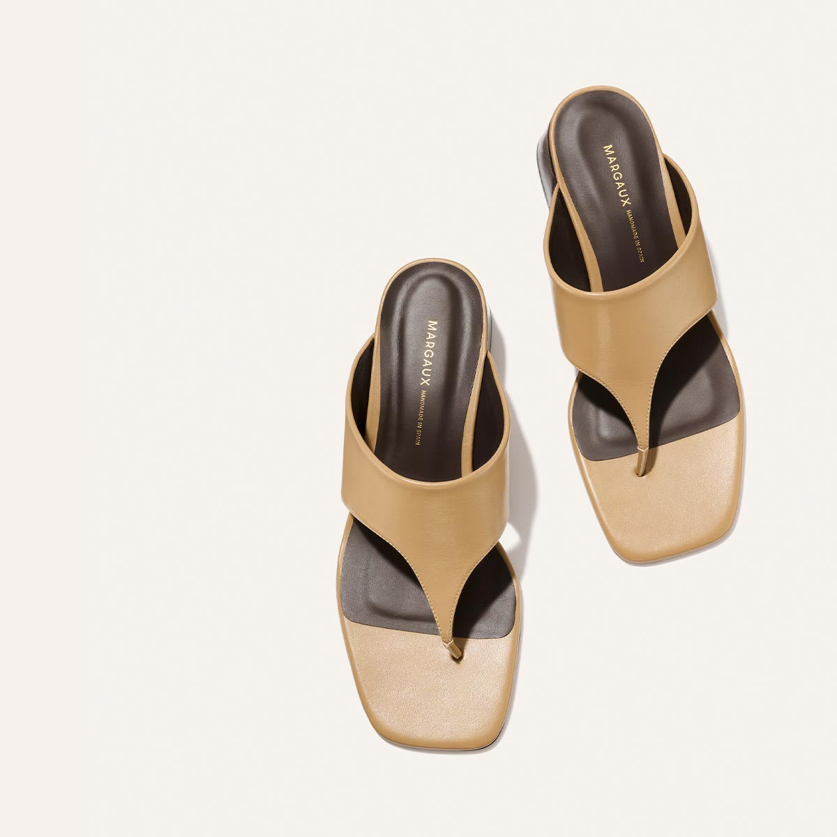 A pair of The Deia 50 - Praline Nappa thong sandals by Margaux, featuring square toes, tan leather straps, black insoles, and gold “Margaux” branding, displayed on a light background.