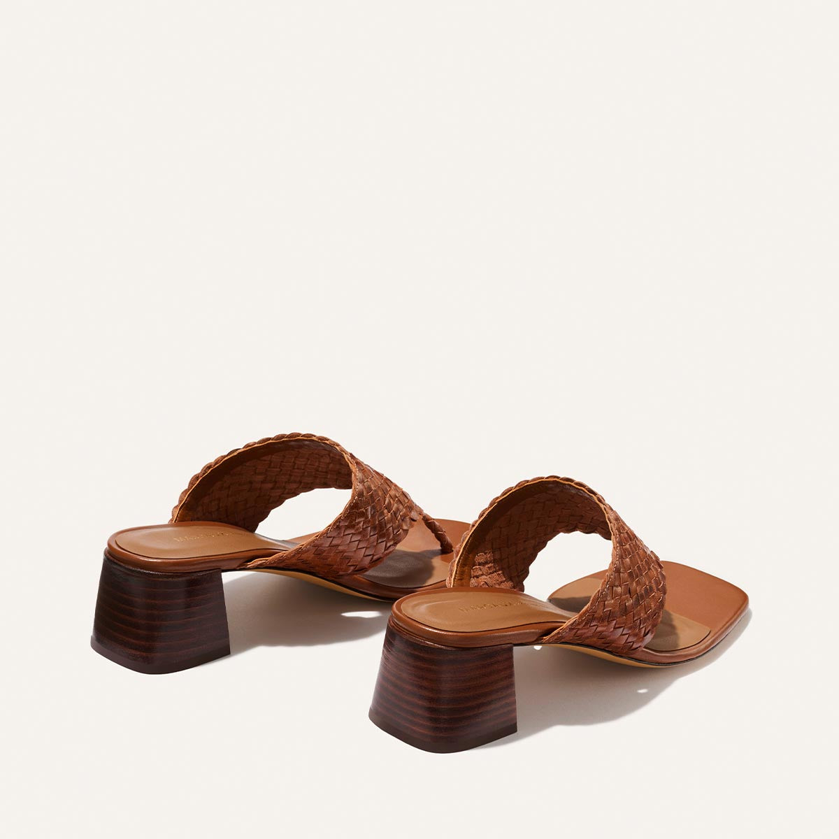 The Deia 50 - Saddle Woven Leather sandals by Margaux feature brown woven leather, a wide foot strap, and medium block heels, shown from the back and side on a plain white background.