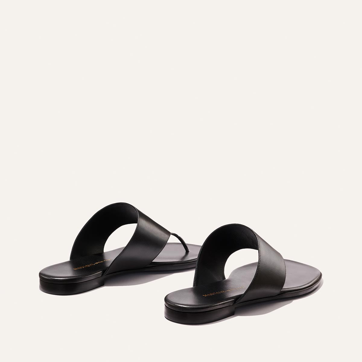The Deia - Black Nappa slip-on sandals by Margaux feature wide black leather straps and flat soles, displayed on a light background with one sandal slightly ahead of the other.