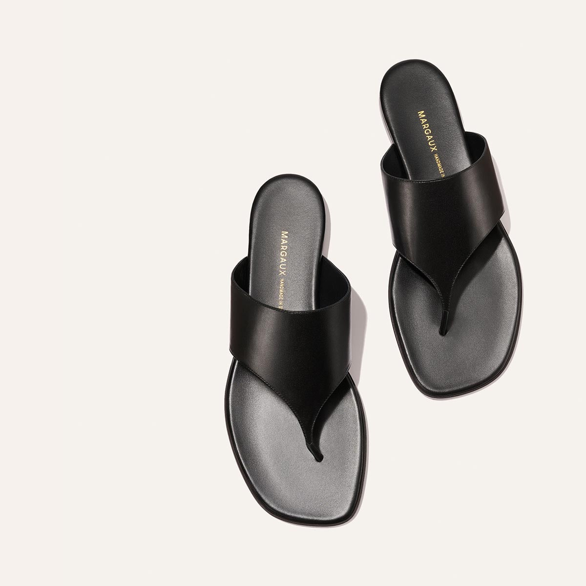 The Deia - Black Nappa by Margaux: minimalist black thong sandals with wide straps, square toes, and gold branding on the insoles, shown on a light background.