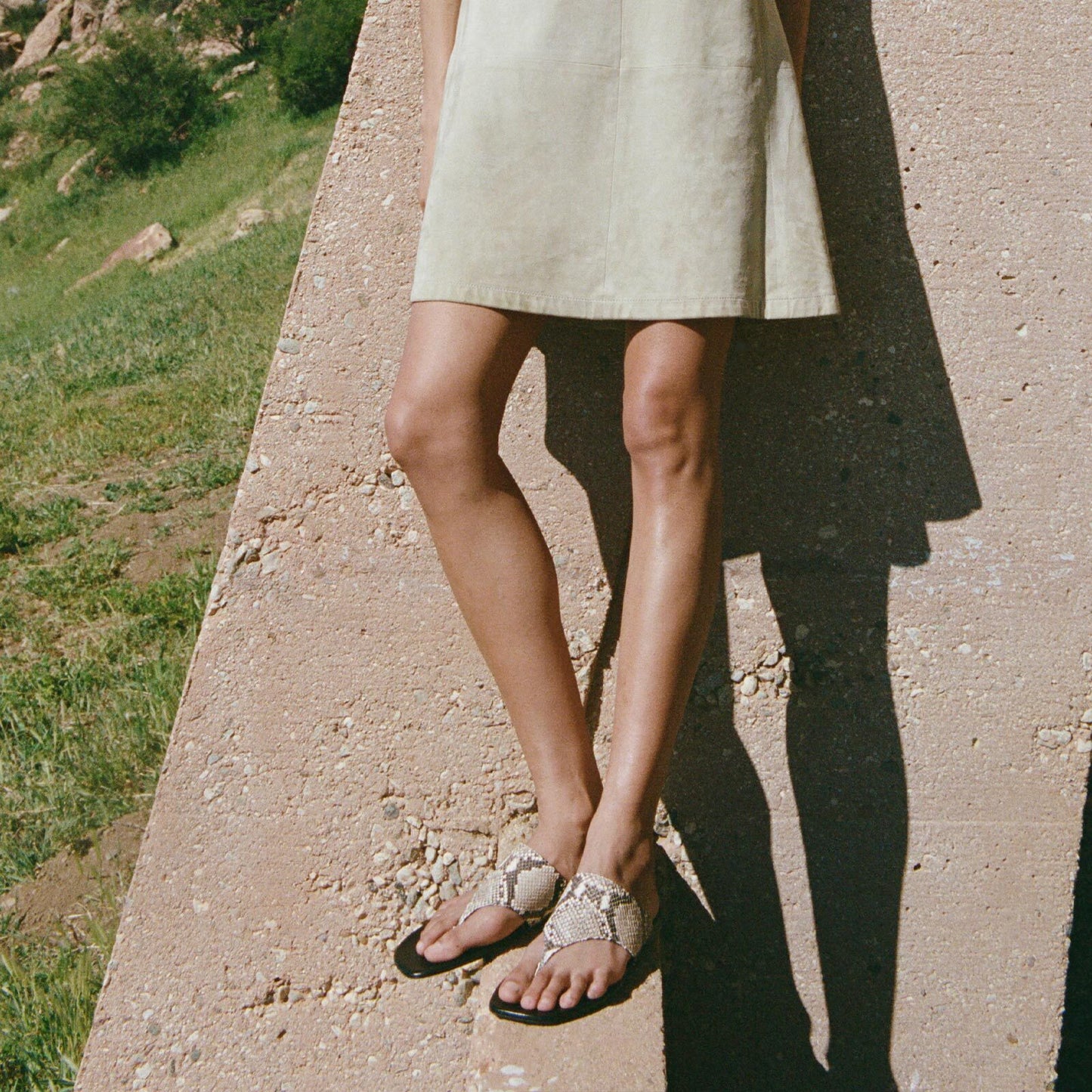 A person wearing a light-colored dress and Margaux's The Deia - Natural Python Embossed sandals stands outdoors by a slanted concrete wall, with grass and rocks in the background.