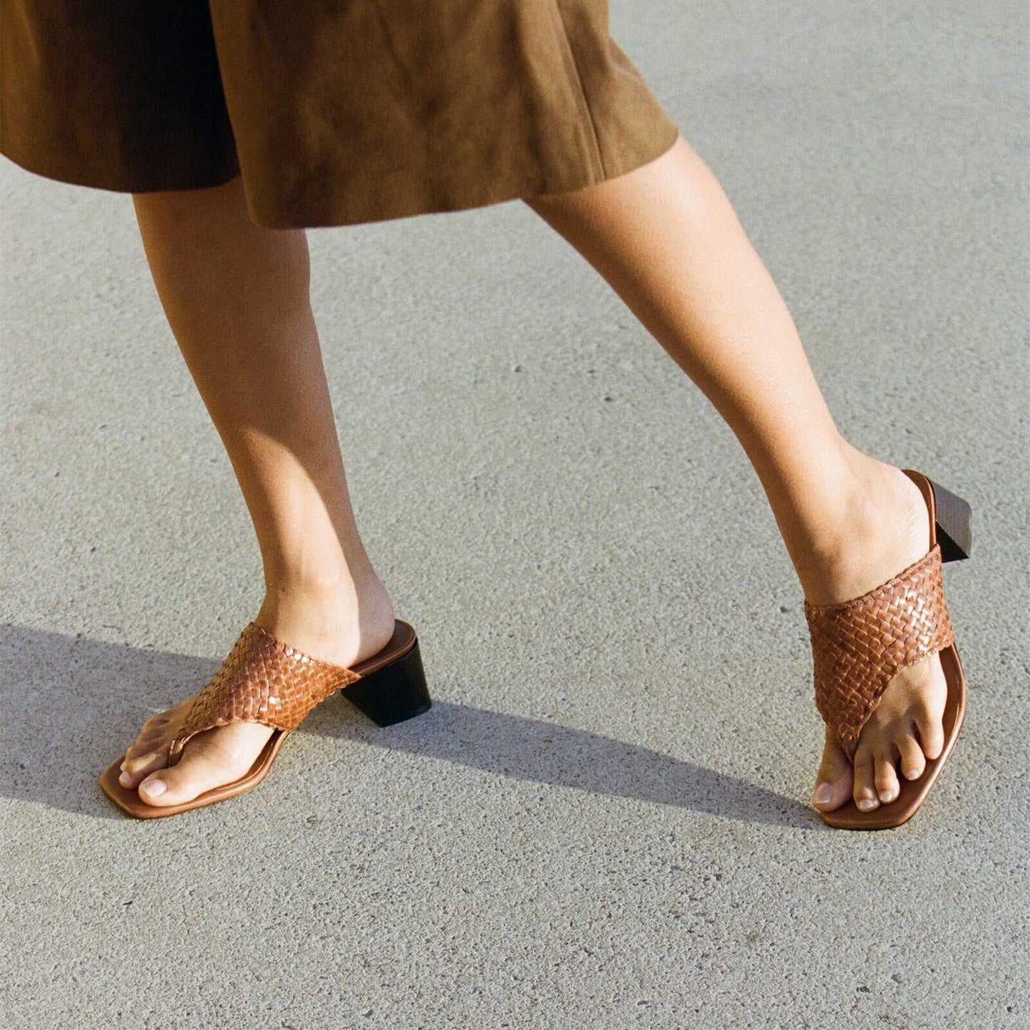 A person in a brown skirt walks on light pavement, wearing Margaux’s The Deia 50 - Saddle Woven Leather heeled sandals that showcase their feet.