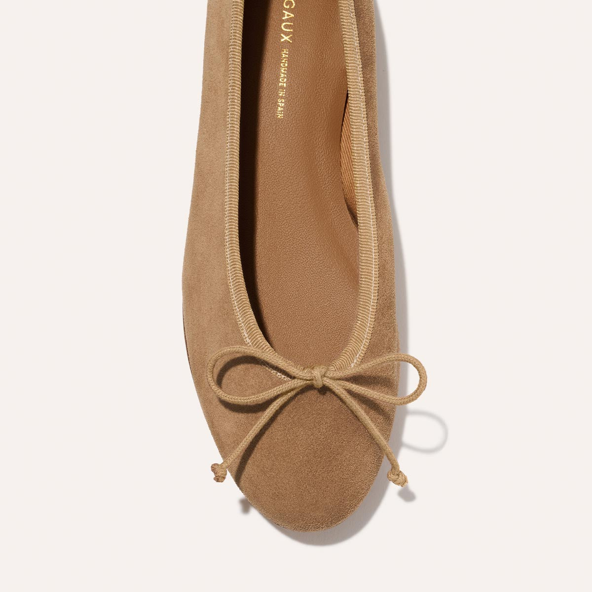 A close-up top view of The Demi - Biscotti Suede by Margaux, a tan suede ballet flat handmade in Spain with a rounded toe, fabric trim, and bow detail at the front.