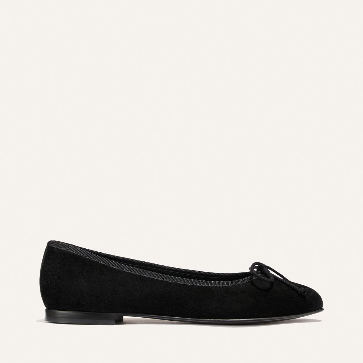 The Demi - Black Suede by margaux-development, a handmade black ballet flat crafted in Spain, features a small bow on the toe and is displayed in a side view against a plain light background.