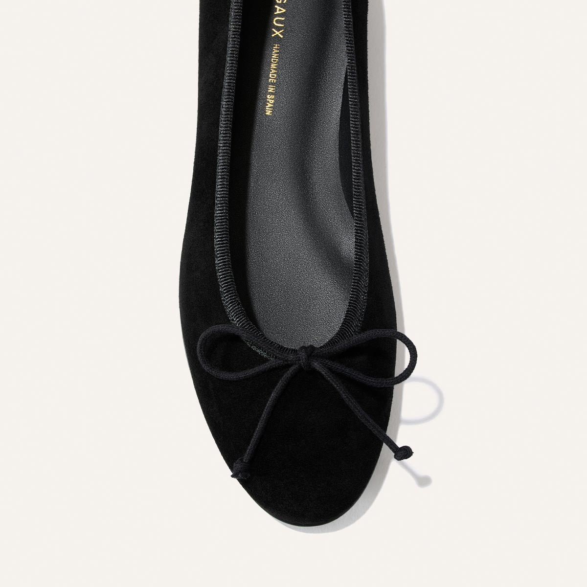 A top view of The Demi - Black Suede ballet flat by margaux-development, handmade in Spain with a rounded toe and a simple bow detail on the front, set against a light background.