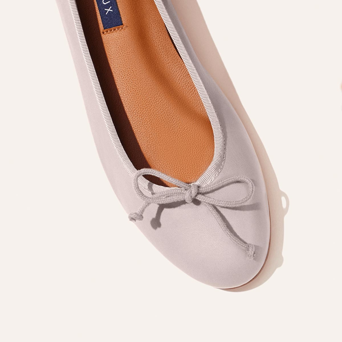 The Demi - Dove Nappa by margaux-development is a light pink ballet flat handmade in Spain, featuring a rounded toe, white trim, front bow, and tan insole—photographed on a neutral background for timeless style and comfort.