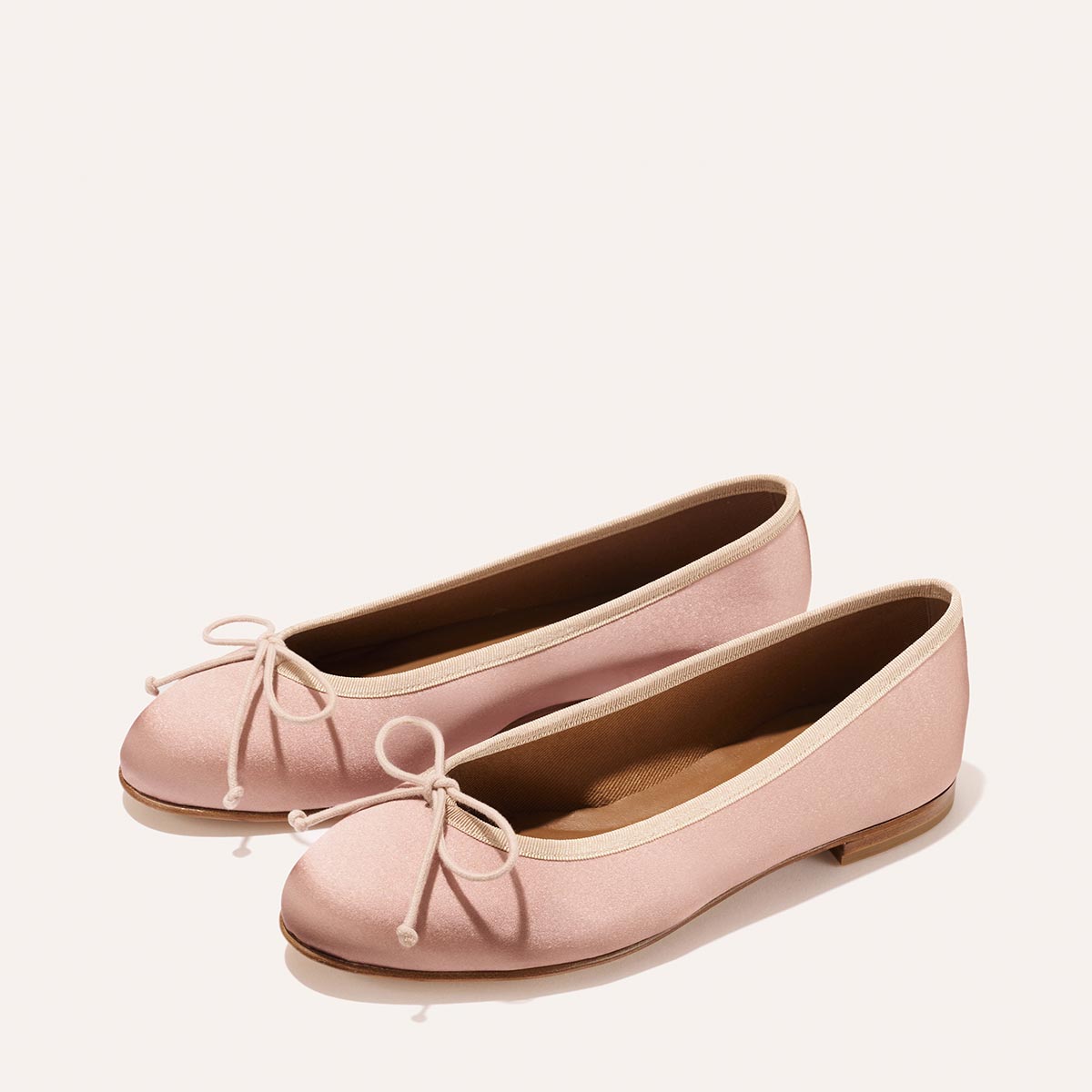 The Demi - Dusty Pink Satin ballet flats by margaux-development, handmade in Spain, feature rounded toes, matching front bows, and light brown soles—shown on a plain off-white background.