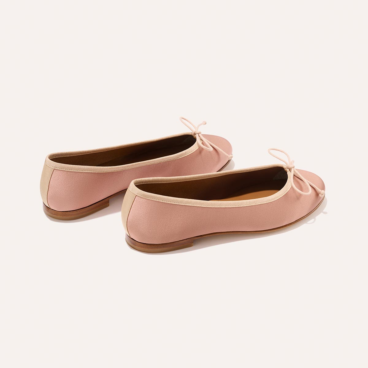 The Demi - Dusty Pink Satin by margaux-development are handmade in Spain: light pink ballet flats with beige trim, small bows on the toes, and brown soles, shown on an off-white background.