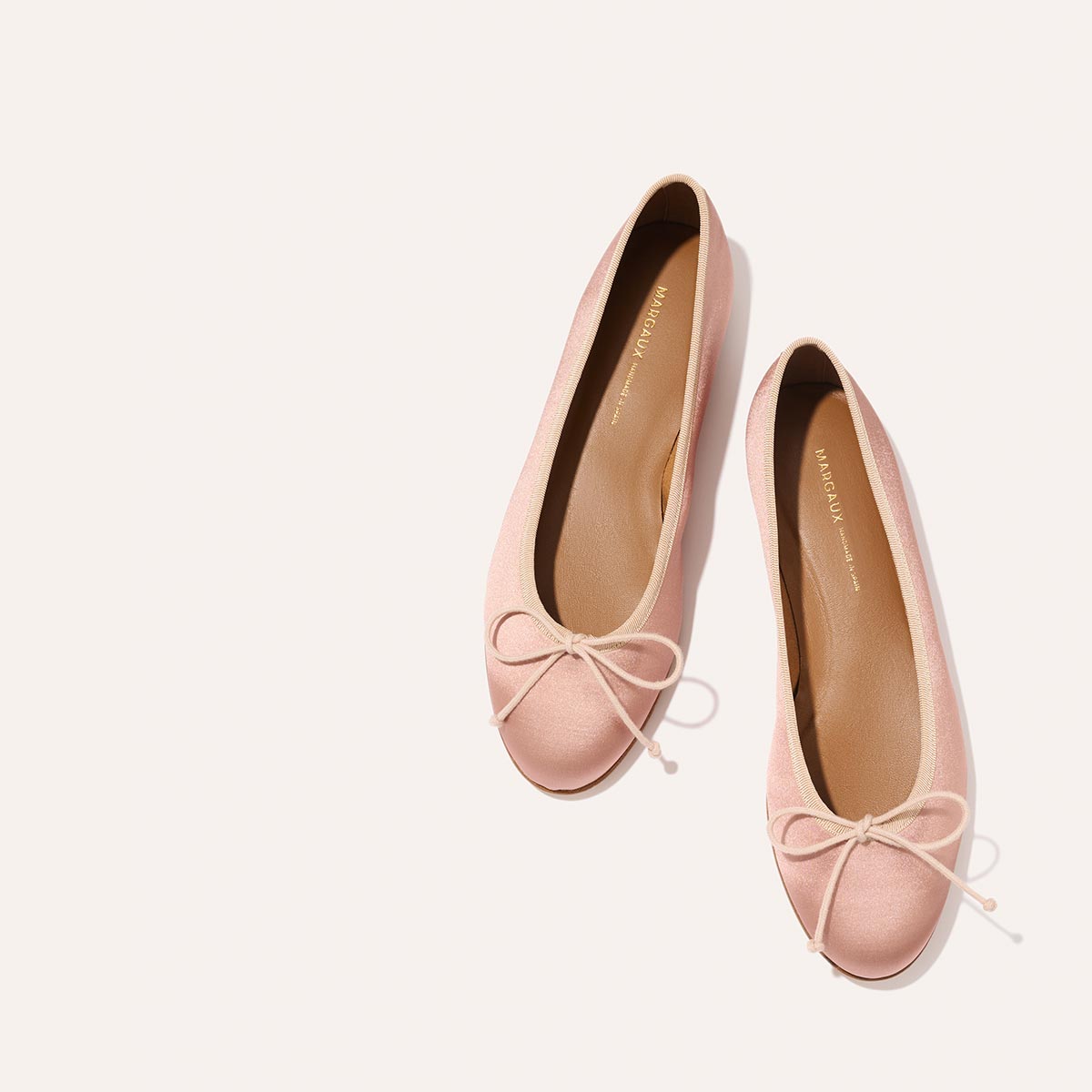 The Demi - Dusty Pink Satin by margaux-development: handmade in Spain, these light pink ballet flats feature small bows on the toes and are shown against a plain, light background.