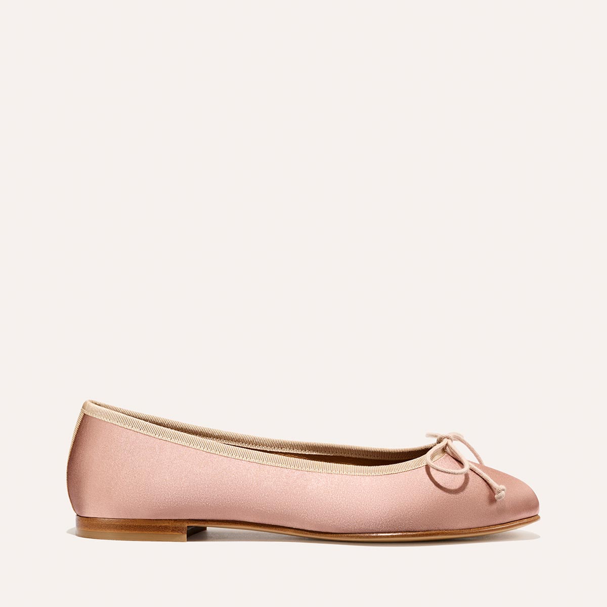 The Demi - Dusty Pink Satin by margaux-development is a handmade light pink ballet flat from Spain, featuring a small bow, beige trim, and a flat wooden sole, showcased on a simple light background.