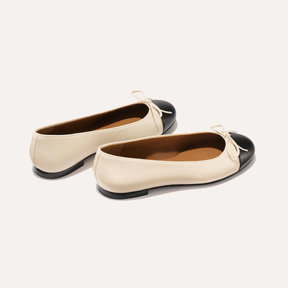 The Demi - Ecru and Black Nappa by margaux-development: cream ballet flats with black rounded toe caps and small front bows, handmade in Spain, shown on a neutral background.