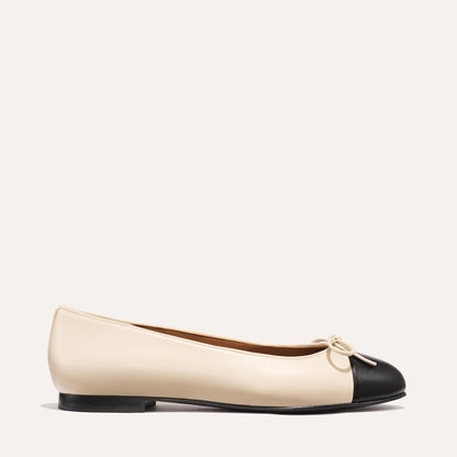 The Demi - Ecru and Black Nappa by margaux-development is a handmade Spanish ballet flat in beige with a black rounded toe cap, small front bow, and low black heel, shown in side profile against a plain light background.