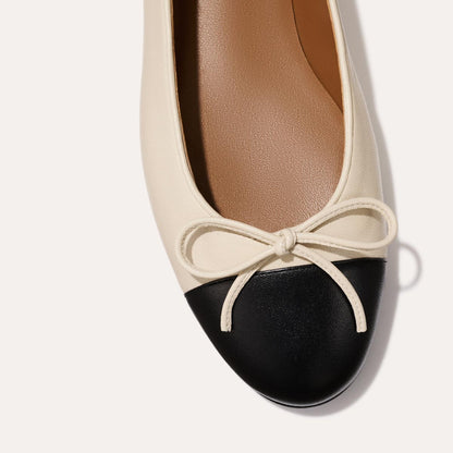 A close-up of The Demi - Ecru and Black Nappa by margaux-development, a bestselling ballet flat handmade in Spain, with an ecru body, black nappa toe cap, and front bow, displayed against a light background.