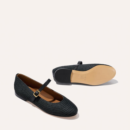 The Demi Jane - Black Raffia by Margaux: a pair of black woven ballet flats featuring a single strap with a gold buckle. One shoe stands upright, the other lies on its side to reveal the light tan sole.