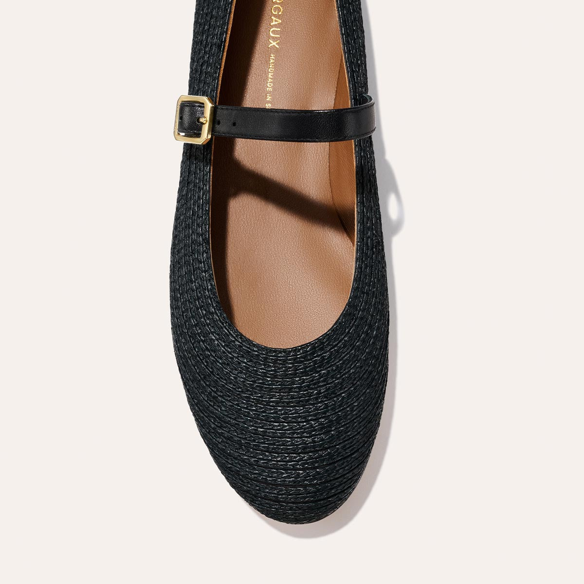 A close-up top view of Margaux's The Demi Jane - Black Raffia reveals a black woven flat with a rounded toe, thin black strap with gold buckle, and tan interior.