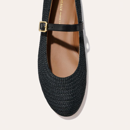 A close-up top view of Margaux's The Demi Jane - Black Raffia reveals a black woven flat with a rounded toe, thin black strap with gold buckle, and tan interior.