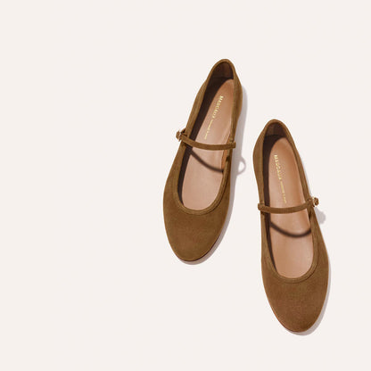 A pair of The Demi Jane - Cedar Suede flats from margaux-development, featuring a single strap and brown suede finish, is displayed on an off-white background with the gold brand name inside, highlighting their elegant femininity.