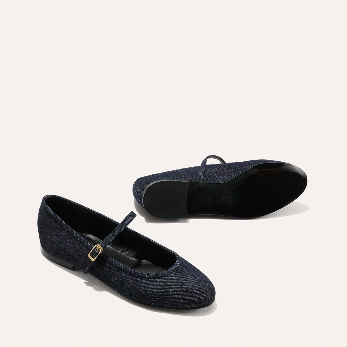 Two Indigo Denim The Demi Jane flats by margaux-development, one standing upright and the other on its side to show the sole, feature a small buckle strap and exude elegant femininity against a plain light background.