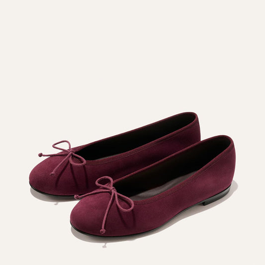 A pair of The Demi - Merlot Suede ballet flats by Margaux, crafted from comfortable burgundy suede with rounded toes and bow accents, handmade in Spain and shown against a plain light background.