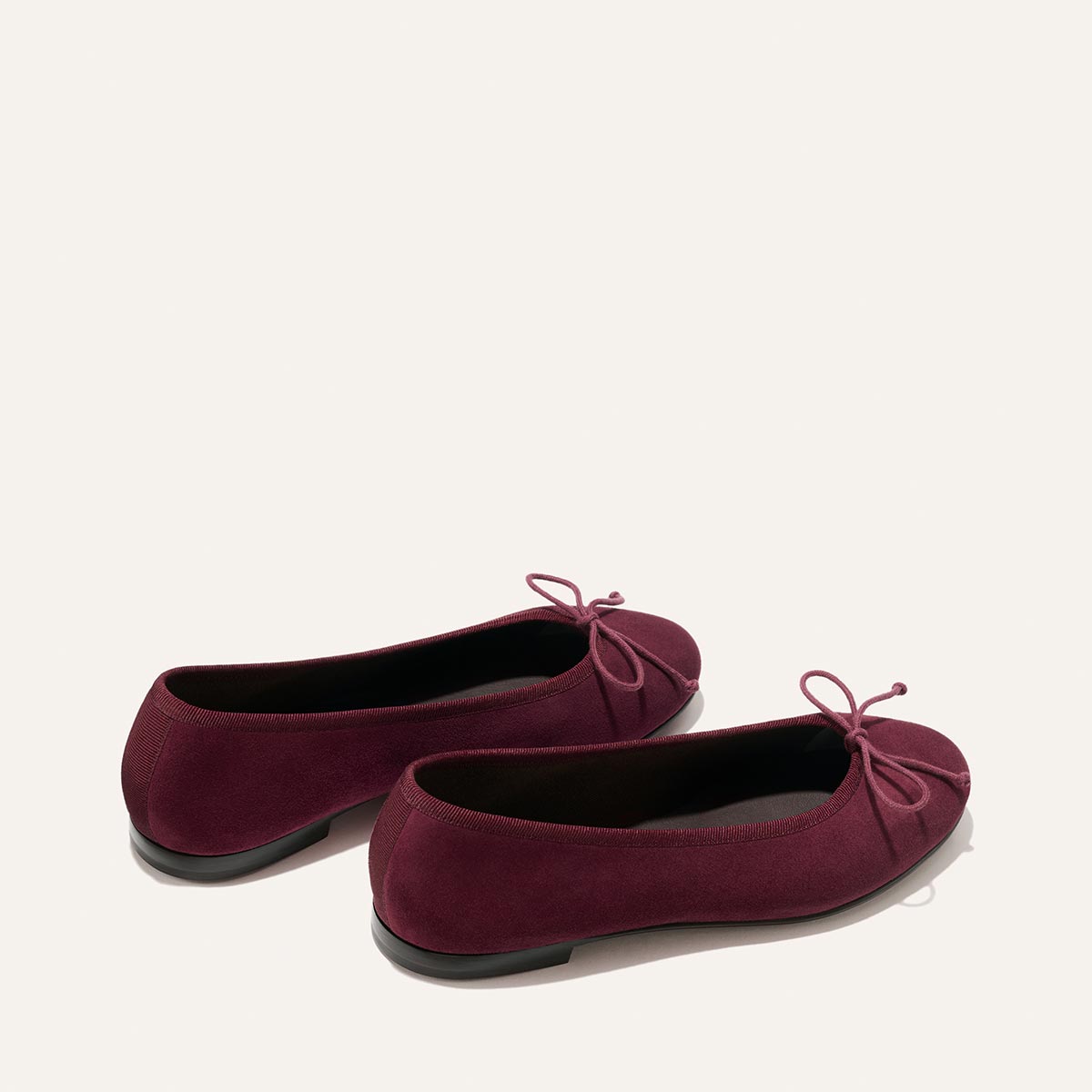 A pair of The Demi - Merlot Suede ballet flats by Margaux, handmade in Spain and adorned with petite bows on the front, are displayed on a neutral background, positioned slightly askew from each other.