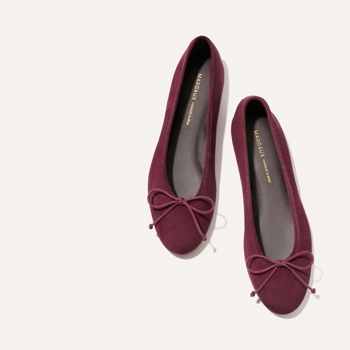 A pair of The Demi - Merlot Suede ballet flats by Margaux, featuring bows on the toes and displayed on a light background. Handmade in Spain, these comfortable flats have gray insoles with “Margaux” printed in gold inside each shoe.