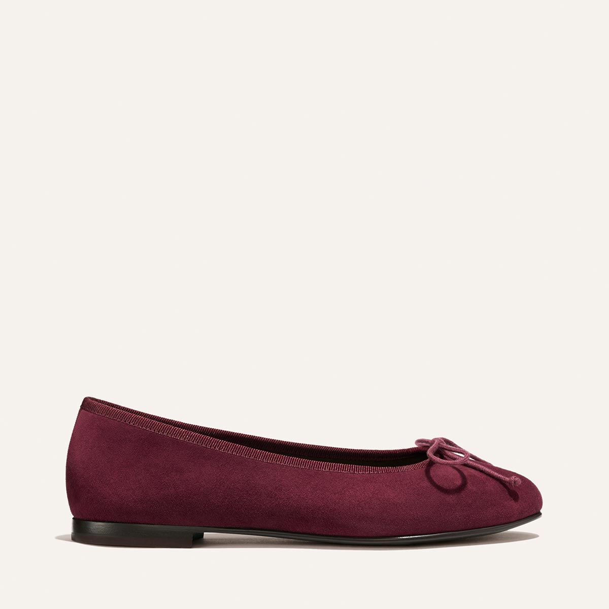 The Demi - Merlot Suede by Margaux is a handmade maroon ballet flat, crafted in Spain with a small bow at the front, shown in side profile against a simple off-white background.