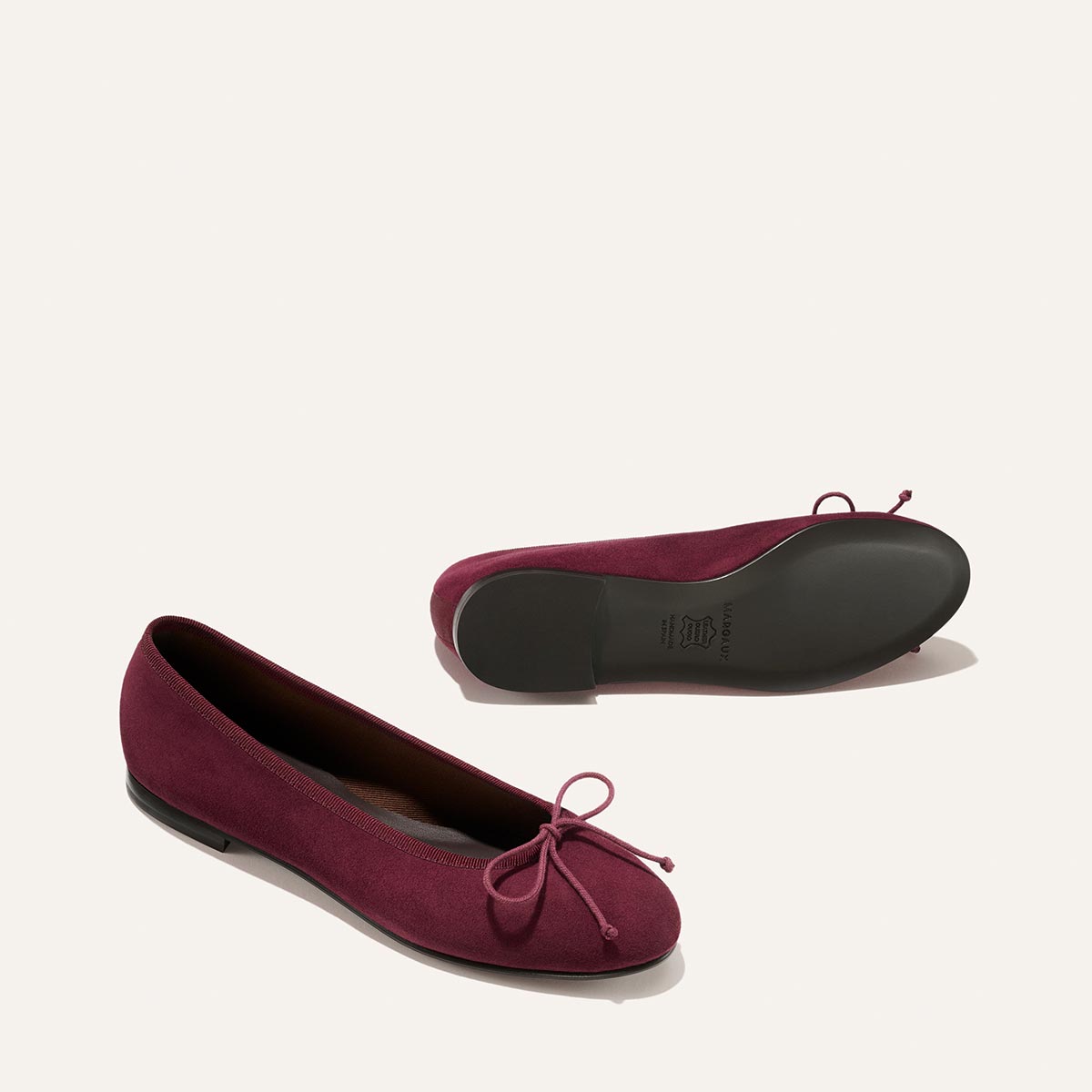 A pair of Margaux’s The Demi - Merlot Suede ballet flats, crafted from soft burgundy suede and handmade in Spain, feature delicate bows on the toes. Photographed against a plain light background, one shoe stands upright while the other rests on its side to showcase the sole.