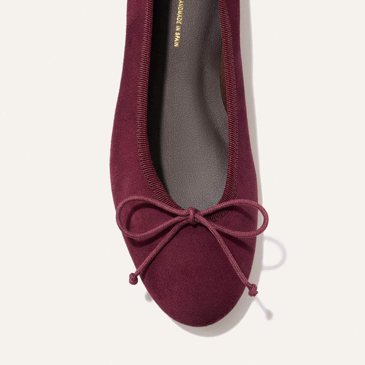 A top view of The Demi - Merlot Suede ballet flat by Margaux, a comfortable burgundy suede shoe featuring a bow at the toe, handmade in Spain, displayed on a light background.