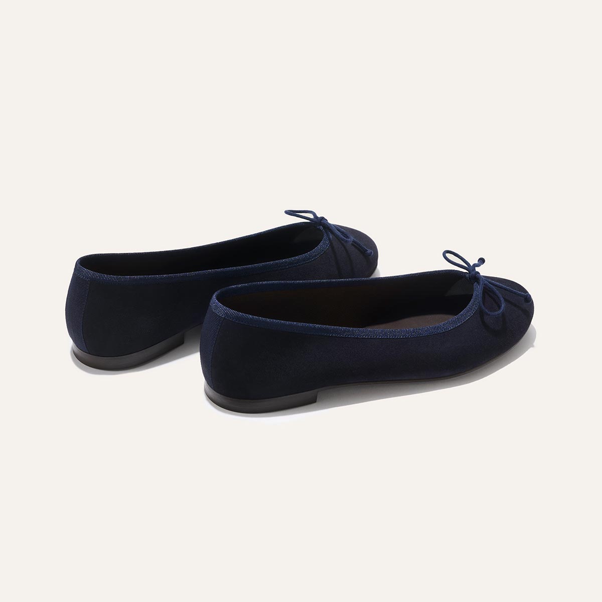 The Demi - Navy Suede by margaux-development: handmade navy blue ballet flats from Spain, featuring small bows on the toes, shown from a rear side angle on a plain light background.
