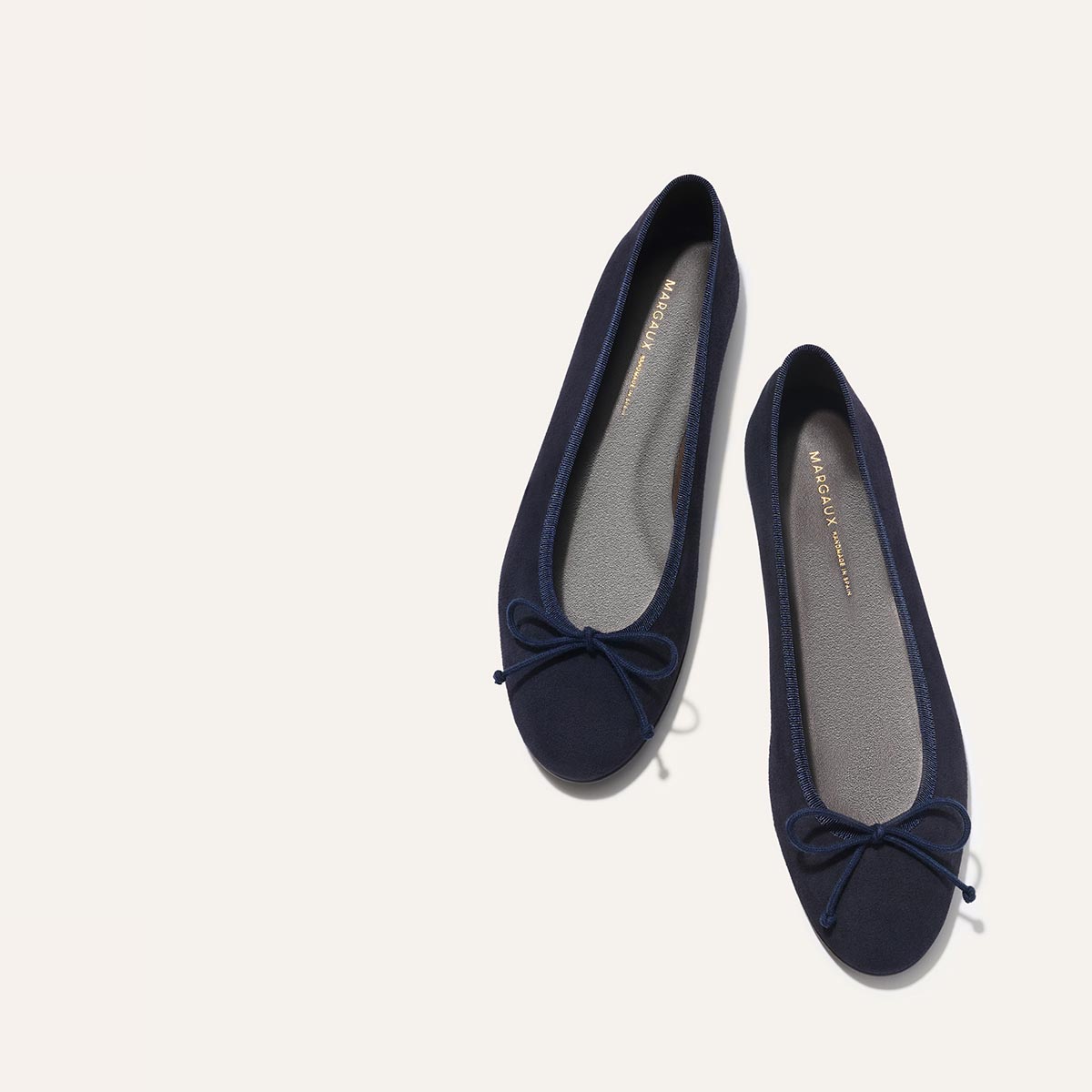 A pair of The Demi - Navy Suede ballet flats by margaux-development, handmade in Spain with small toe bows, displayed side by side on a light off-white background.