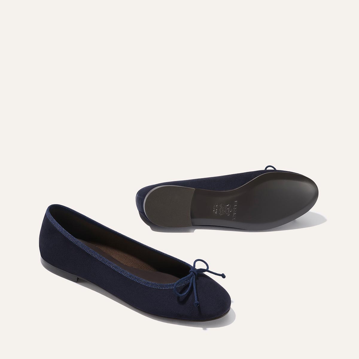 The Demi - Navy Suede by margaux-development are handmade in Spain. These navy ballet flats feature small front bows; one stands upright and the other lies on its side to highlight the sole against a light background.