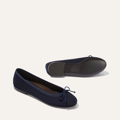 The Demi - Navy Suede by margaux-development are handmade in Spain. These navy ballet flats feature small front bows; one stands upright and the other lies on its side to highlight the sole against a light background.