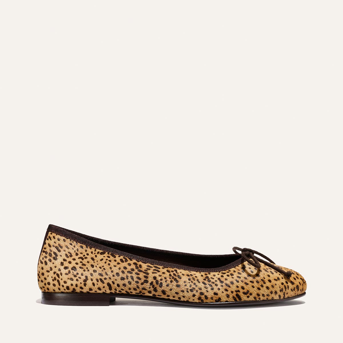 A single Margaux “The Demi - Tan Mini Cheetah Haircalf” ballet flat, handmade in Spain, featuring cheetah-print haircalf with a small bow on the toe and black trim, is shown in profile against a plain light background.