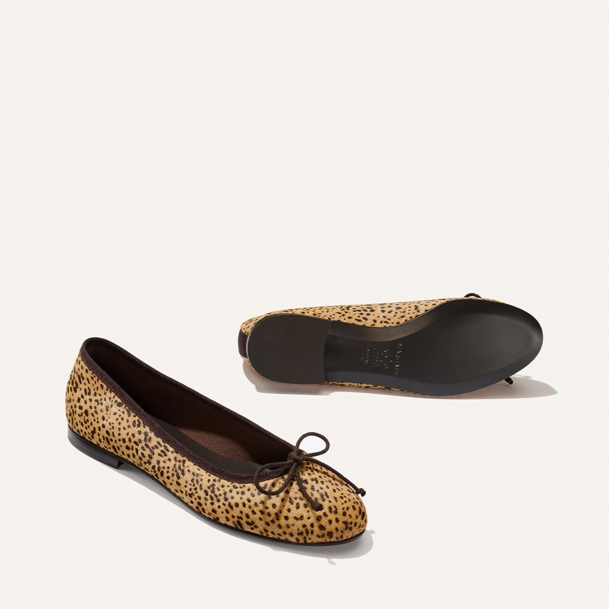 Two “The Demi - Tan Mini Cheetah Haircalf” ballet flats by Margaux—featuring a chic cheetah-print haircalf exterior, black trim, and delicate bows—are displayed with one shoe standing upright and the other resting on its side to showcase the sole. Handmade in Spain, these comfortable flats combine signature Margaux style with expert craftsmanship.
