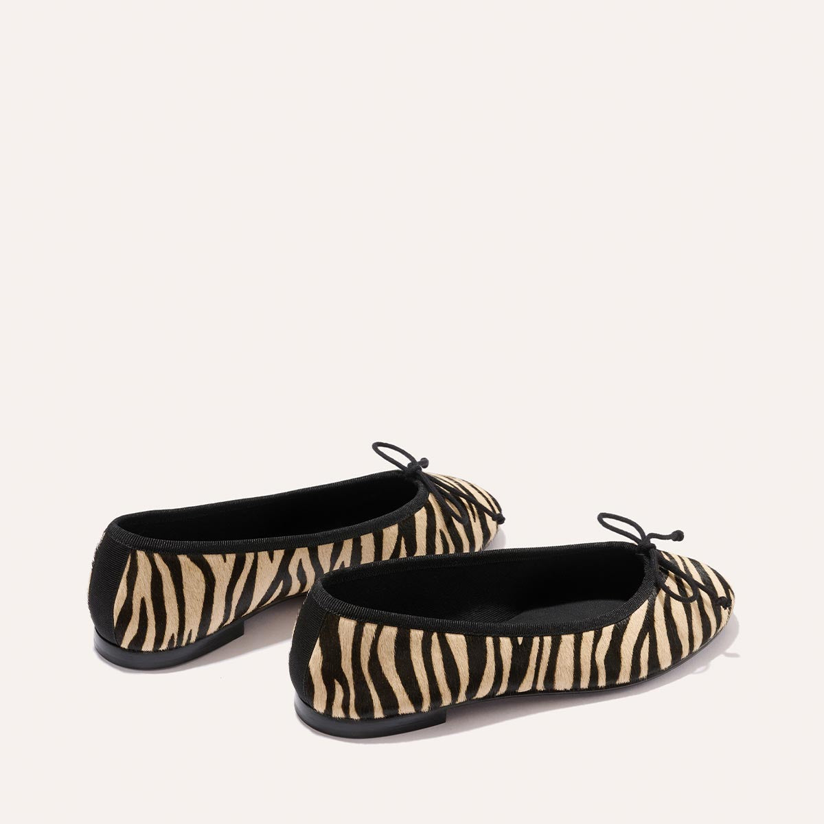 The Margaux Demi - Tan Zebra Haircalf: stylish zebra-print ballet flats with black trim, black toe bows, and flat black soles, shown on a light beige background.
