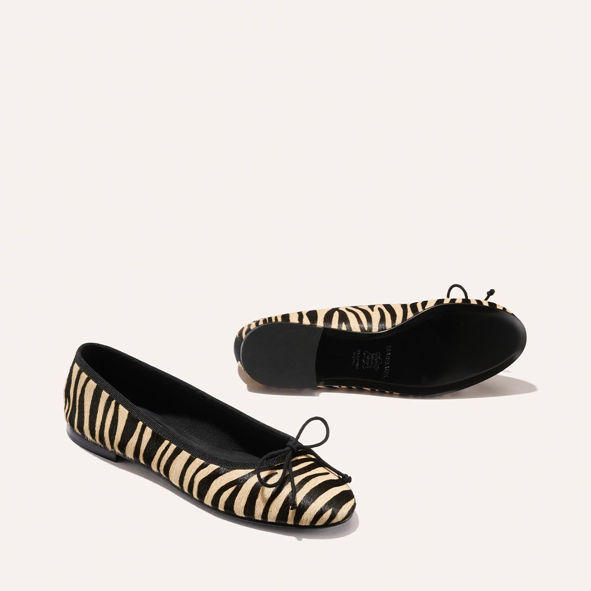 The Demi - Tan Zebra Haircalf by Margaux: handmade in Spain, these comfortable ballet flats feature black and beige zebra stripes, black trim, and small black bows at the toes, shown on a light background.