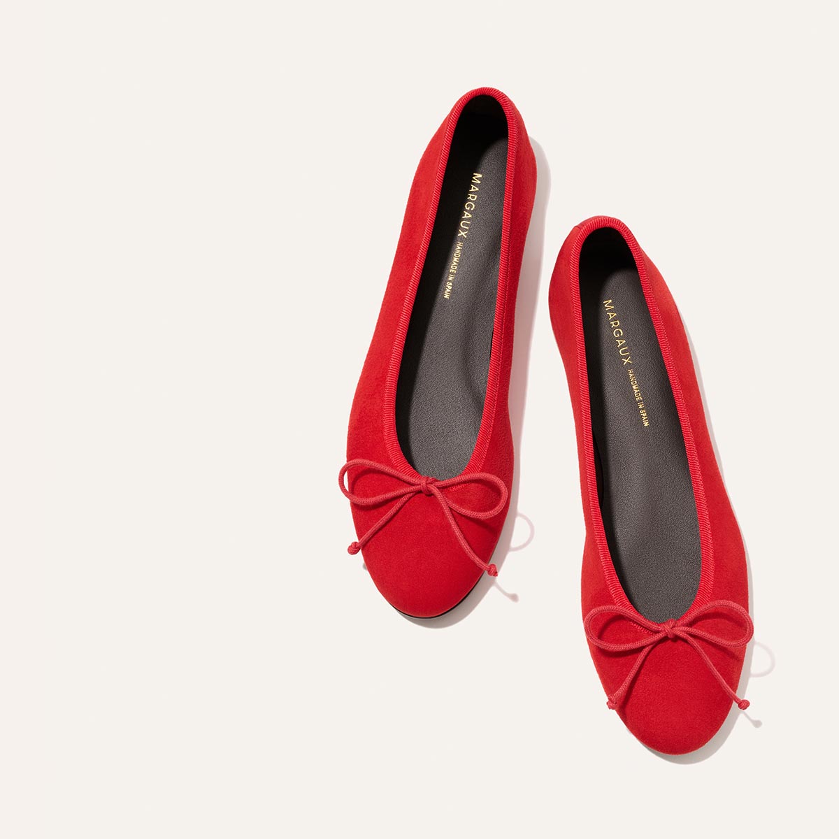 Two The Demi - Tomato Suede ballet flats by Margaux, featuring bow details, are displayed side by side on a white background. Handmade in Spain with black insoles and gold branding.
