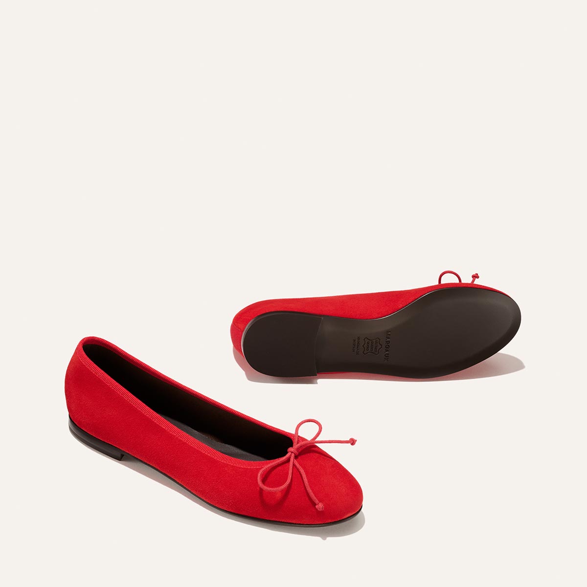 Two Tomato Suede The Demi flats by Margaux—one upright showing the side and bow, the other on its side revealing the sole—are set against a light cream background.