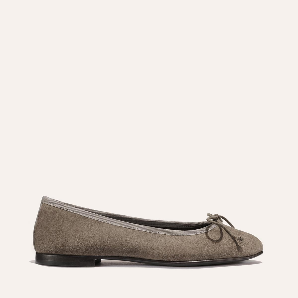 The Demi - Truffle Suede by Margaux is a comfortable ballet flat, handmade in Spain from taupe suede, featuring a black sole, gray trim around the opening, and a delicate bow on the toe—shown in side profile against a plain light background.