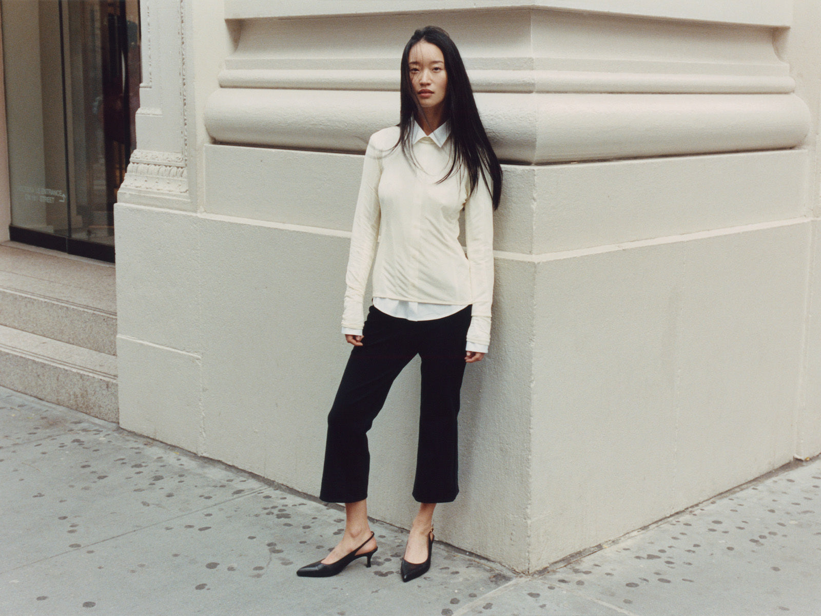 Woman in a white blouse and black pants standing against a light-colored building.