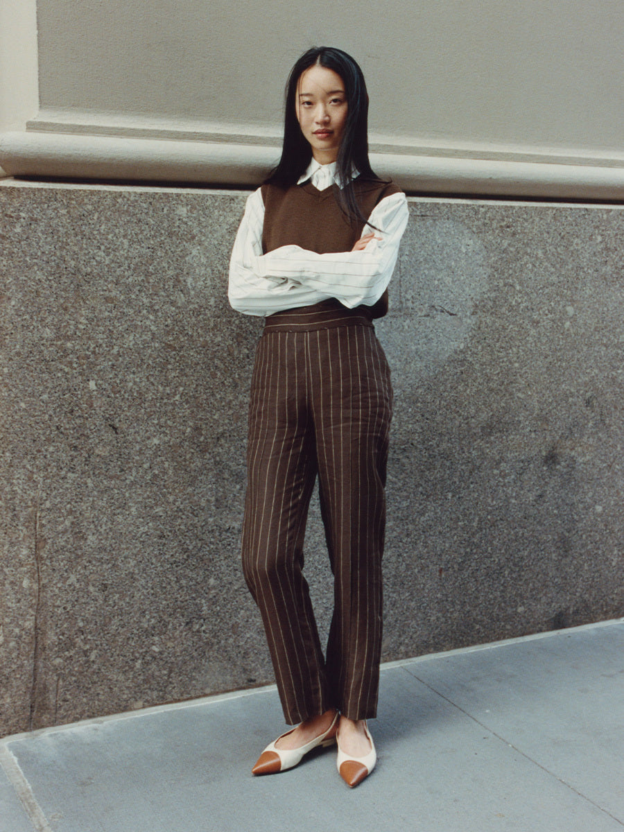 Woman wearing a brown vest and striped pants standing against a gray wall.