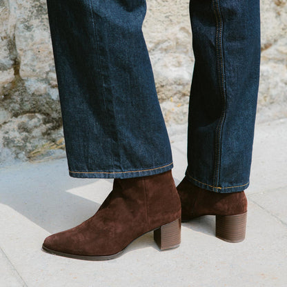 A person wearing dark blue denim jeans and Margauxs The Downtown Boot in Chocolate Suede v1 stands on light pavement with a stone wall behind them.