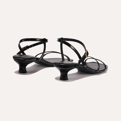 The Dylan Sandal in shiny Black Crinkle-Gloss Nappa, featuring asymmetrical straps around the ankle and toes and a walkable 1-inch stiletto heel.