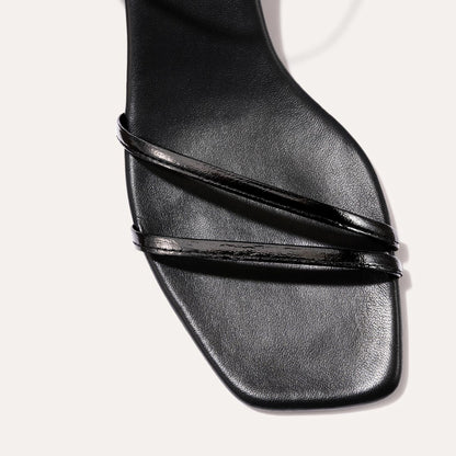 The Dylan Sandal in shiny Black Crinkle-Gloss Nappa, featuring asymmetrical straps around the ankle and toes and a walkable 1-inch stiletto heel.