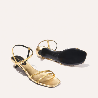 The Dylan Sandal - Gold Mirror by margaux-development: a pair of gold strappy sandals with low heels. One sandal stands upright showing straps and ankle buckle; the other is flipped to reveal the black sole, all on an off-white background.